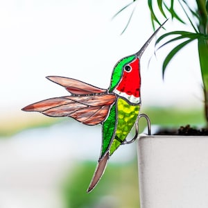 Könnte beinhalten: Ein bunter Glas-Kolibri mit roten, grünen und braunen Akzenten sitzt auf einem Metallhaken. Der Kolibri ist nach rechts gerichtet und hat seine Flügel ausgebreitet. Der Haken ist an einem weißen Topf befestigt.