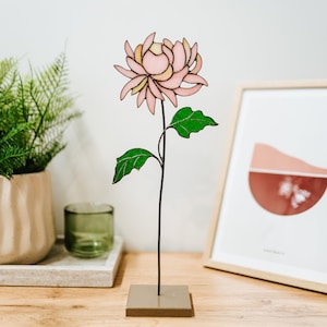 May include: A pink stained glass flower with green leaves on a black metal stem. The flower is standing on a square gray base.