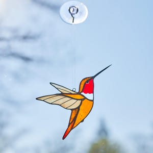 K&ouml;nnte beinhalten: Ein Glasfenster-Kolibri-Sonnenf&auml;nger mit rotem Hals, orangefarbenem K&ouml;rper und beigefarbenen Fl&uuml;geln. Der Vogel ist an einem transparenten Saugnapf aufgeh&auml;ngt. Der Kolibri ist ein dekorativer Artikel.