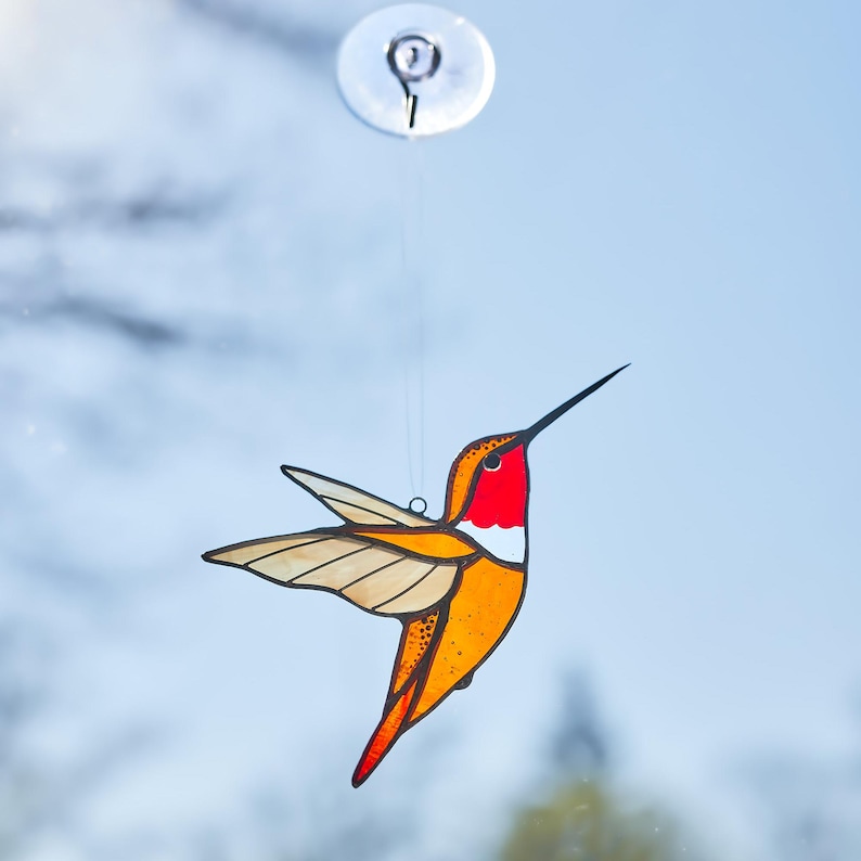 K&ouml;nnte beinhalten: Ein Glasfenster-Kolibri-Sonnenf&auml;nger mit rotem Hals, orangefarbenem K&ouml;rper und beigefarbenen Fl&uuml;geln. Der Vogel ist an einem transparenten Saugnapf aufgeh&auml;ngt. Der Kolibri ist ein dekorativer Artikel.