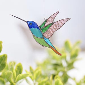 K&ouml;nnte beinhalten: Ein Buntglas-Kolibri mit blauen, gr&uuml;nen und orangefarbenen Akzenten. Der Kolibri ist im Flug mit ausgebreiteten Fl&uuml;geln.