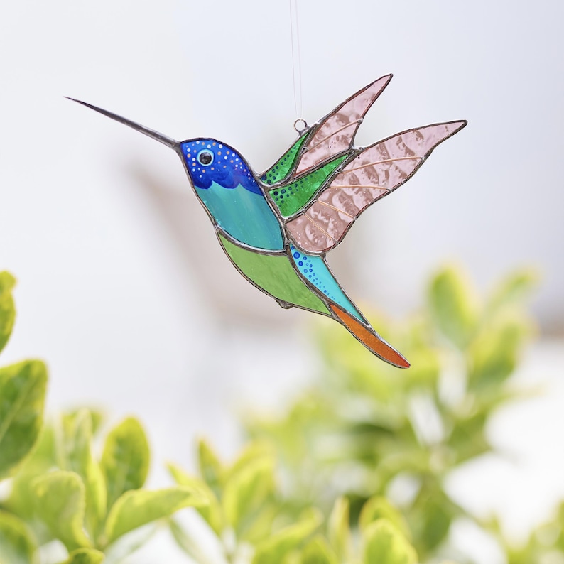 K&ouml;nnte beinhalten: Ein Buntglas-Kolibri mit blauen, gr&uuml;nen und orangefarbenen Akzenten. Der Kolibri ist im Flug mit ausgebreiteten Fl&uuml;geln.