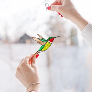 K&ouml;nnte beinhalten: Ein Buntglas-Kolibri mit gr&uuml;nen, gelben und roten Akzenten, der an einem Fenster h&auml;ngt.