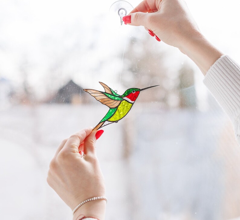 K&ouml;nnte beinhalten: Ein Buntglas-Kolibri mit gr&uuml;nen, gelben und roten Akzenten, der an einem Fenster h&auml;ngt.