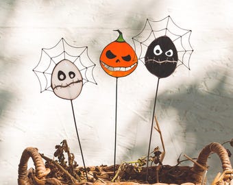 May include: Three Halloween-themed glass ornaments with wire spiderwebs. The ornaments are a pumpkin with a scary face, a white ghost with a stitched mouth, and a black ghost with a stitched mouth.