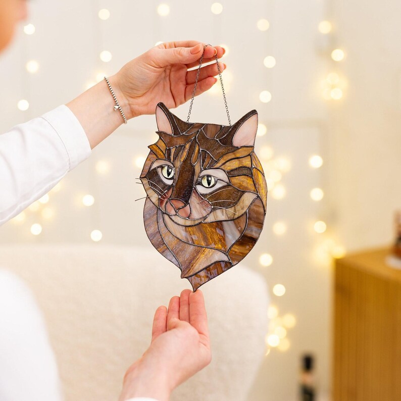 May include: A stained glass cat head hanging from a silver chain. The cat has brown, black, and white fur and green eyes.