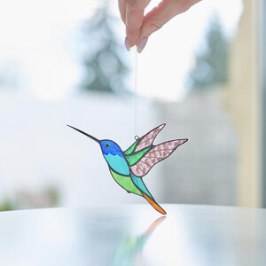 K&ouml;nnte beinhalten: Ein Ornament aus Buntglas in Form eines Kolibris mit blauen, gr&uuml;nen und rosa Akzenten. Der Kolibri ist im Flug mit ausgebreiteten Fl&uuml;geln. Das Ornament h&auml;ngt an einer Schnur.