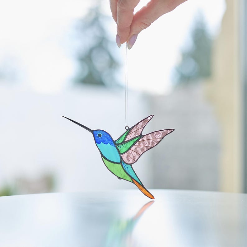 K&ouml;nnte beinhalten: Ein Ornament aus Buntglas in Form eines Kolibris mit blauen, gr&uuml;nen und rosa Akzenten. Der Kolibri ist im Flug mit ausgebreiteten Fl&uuml;geln. Das Ornament h&auml;ngt an einer Schnur.