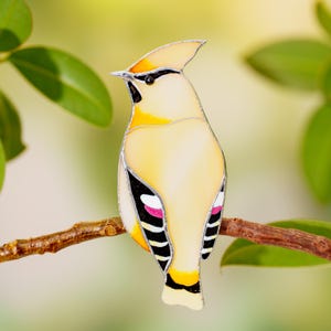 May include: A stained glass bird with a yellow body, black and white wing accents, and a crest. The bird is perched on a brown branch against a blurred green and yellow background. The bird's beak is black, and the wings have pink accents.