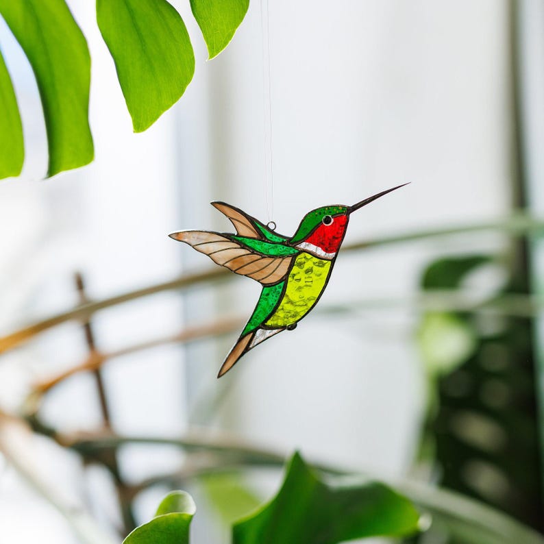 K&ouml;nnte beinhalten: Ein Buntglas-Kolibri mit rotem Kopf, gr&uuml;nem K&ouml;rper und gelben Akzenten. Der Kolibri h&auml;ngt an einer Schnur.