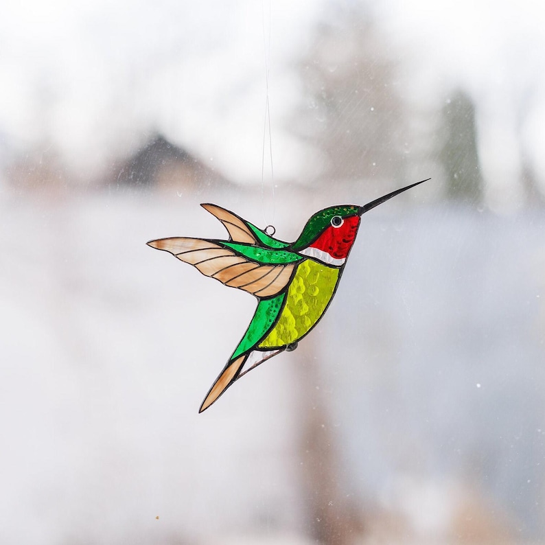 K&ouml;nnte beinhalten: Ein Buntglas-Kolibri mit gr&uuml;nen, gelben, roten und braunen Akzenten. Der Kolibri ist im Flug mit ausgebreiteten Fl&uuml;geln.