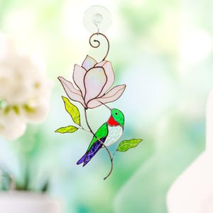 May include: A stained glass hummingbird with a green head, red throat, and blue and white body, near a light pink flower. The artwork includes green leaves and a brown stem, attached to a window with a suction cup.