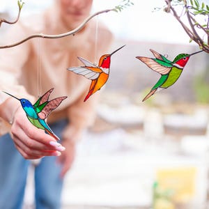 K&ouml;nnte beinhalten: Drei Kolibri-Ornamente aus Buntglas, die an einem Ast h&auml;ngen. Die Kolibris sind orange, gr&uuml;n und blau mit roten Akzenten.
