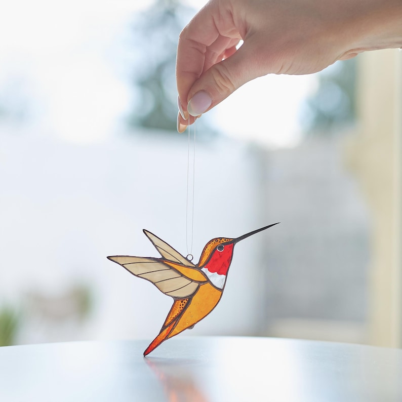 K&ouml;nnte beinhalten: Ein Glasmosaik-Kolibri-Anh&auml;nger mit rotem Kopf, gelbem K&ouml;rper und braunen Fl&uuml;geln. Der Anh&auml;nger h&auml;ngt an einer wei&szlig;en Schnur.