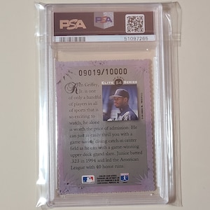 May include: A PSA graded baseball card featuring Ken Griffey Jr. in a white uniform with a black hat. The card is numbered 09019/10000 and is from the Elite 54 Series. The card has a purple and silver background with a white border. The text on the card reads "Ken Griffey, Jr. is one of only a handful of players in all of sports that is so exciting to watch, he alone is worth the price of admission. He can just as easily thrill you with a game-saving diving catch in center field as he can with a game-winning upper deck grand slam. Junior batted .323 in 1994 and led the American League with 40 home runs."
