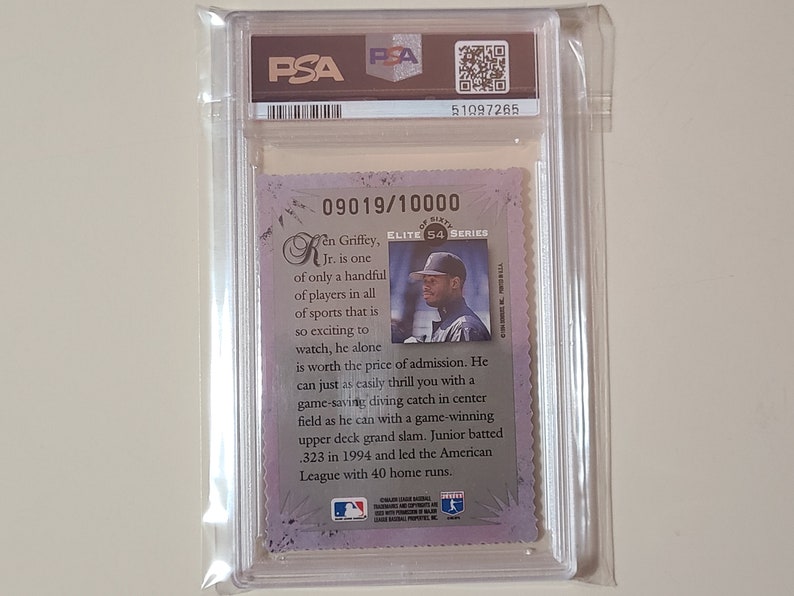 May include: A PSA graded baseball card featuring Ken Griffey Jr. in a white uniform with a black hat. The card is numbered 09019/10000 and is from the Elite 54 Series. The card has a purple and silver background with a white border. The text on the card reads "Ken Griffey, Jr. is one of only a handful of players in all of sports that is so exciting to watch, he alone is worth the price of admission. He can just as easily thrill you with a game-saving diving catch in center field as he can with a game-winning upper deck grand slam. Junior batted .323 in 1994 and led the American League with 40 home runs."