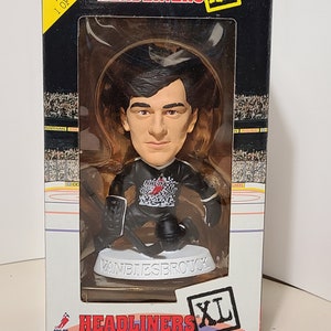 May include: A limited edition Headliners XL bobblehead of a hockey player wearing a black jersey with a red and white logo. The bobblehead is in a clear plastic case on a white base with the player's name, "Vanbiesbrouck", printed on it. The box is black with white and red accents and says "Headliners XL" and "You'll know them when you see them."