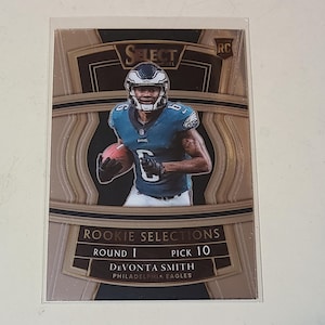 May include: A trading card featuring Devonta Smith of the Philadelphia Eagles. The card has a gold and brown design with the text "Select" at the top. The card also includes the text "Rookie Selections" and "Round 1 Pick 10".