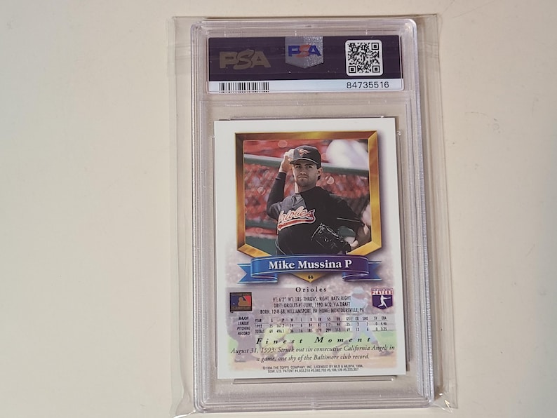 Puede incluir: Una tarjeta de b&eacute;isbol de Mike Mussina certificada por PSA en un estuche de pl&aacute;stico transparente. La tarjeta muestra una fotograf&iacute;a del lanzador con un uniforme negro de los Orioles, con el texto Mike Mussina P, Orioles y Finest Moment.