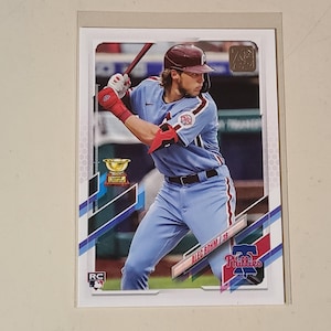 May include: A Topps baseball trading card featuring Alec Bohm of the Philadelphia Phillies in a light blue uniform. The player is shown in a batting stance wearing a maroon helmet and red batting gloves. The card includes a gold trophy icon and RC logo.