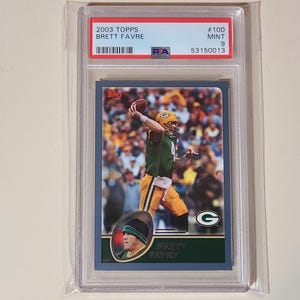 May include: A 2003 Topps football trading card featuring Brett Favre in a Green Bay Packers uniform. The card is graded and encased in a protective holder. The card has a blue border and includes the Packers' logo.