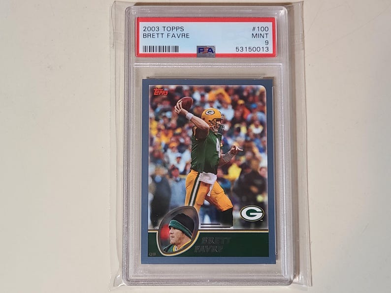 May include: A 2003 Topps football trading card featuring Brett Favre in a Green Bay Packers uniform. The card is graded and encased in a protective holder. The card has a blue border and includes the Packers' logo.