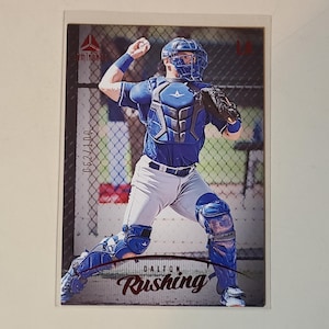 May include: Baseball card featuring a baseball catcher in a blue and white uniform, poised to throw the ball. The card has the text "Dalton Rushing" and "062/100" printed on it.