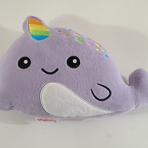 May include: A plush, lavender narwhal toy with a rainbow horn and colorful spots. The toy has a smiling face with black eyes and a white belly. The tail is curved, and the toy is designed for children.
