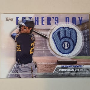May include: A Topps baseball card featuring Christian Yelich of the Milwaukee Brewers. The card has a Father's Day theme with a patch of a baseball glove. Yelich is shown in his Brewers uniform, swinging a bat.