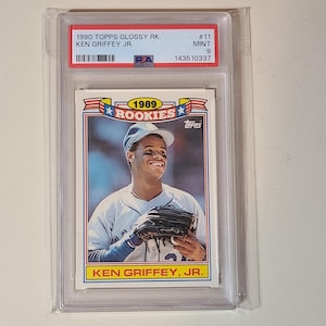 Peut inclure: Une carte de baseball 1990 Topps Glossy Rookies Ken Griffey Jr., not&eacute;e PSA Mint 9. La carte pr&eacute;sente un portrait du joueur portant une casquette et un gant, avec le texte 1989 ROOKIES et KEN GRIFFEY, JR. imprim&eacute; sur le devant de la carte.