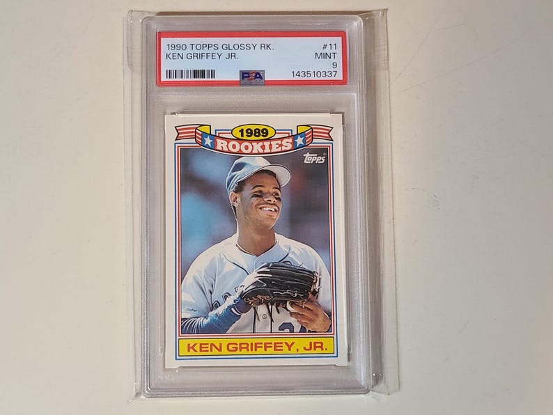 Peut inclure: Une carte de baseball 1990 Topps Glossy Rookies Ken Griffey Jr., not&eacute;e PSA Mint 9. La carte pr&eacute;sente un portrait du joueur portant une casquette et un gant, avec le texte 1989 ROOKIES et KEN GRIFFEY, JR. imprim&eacute; sur le devant de la carte.