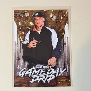 May include: A Topps baseball card featuring Aaron Judge in a black and white jacket and a New York Yankees cap. The card has the text "AARON JUDGE" and "GAMEDAY DRIP" in a stylized font. The background has a gold and brown design.