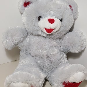 May include: A plush gray teddy bear with a white muzzle and red heart-shaped nose and mouth. The bear has red accents on its ears and feet, with "2019" embroidered on one foot. The bear is sitting upright.