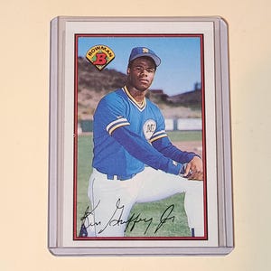 May include: A 1989 Bowman Ken Griffey Jr. baseball card encased in a clear plastic protective sleeve. The card features a portrait of the player in a blue and yellow Seattle Mariners uniform, with the Bowman logo and a printed signature at the bottom.