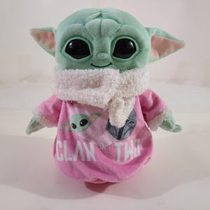 Pu&ograve; includere: Un peluche di un personaggio con grandi occhi neri, pelle verde e grandi orecchie. Il giocattolo indossa un completo rosa con le parole "Clan of Two" e una grafica del personaggio. Una sciarpa bianca &egrave; intorno al collo.