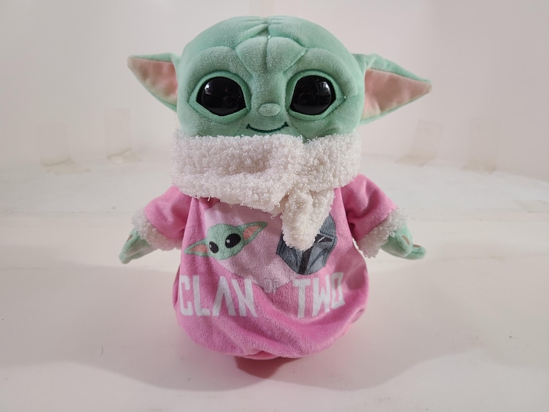 Pu&ograve; includere: Un peluche di un personaggio con grandi occhi neri, pelle verde e grandi orecchie. Il giocattolo indossa un completo rosa con le parole "Clan of Two" e una grafica del personaggio. Una sciarpa bianca &egrave; intorno al collo.
