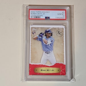 Op de afbeelding: Een honkbalkaart van Bobby Witt Jr. in een Kansas City Royals-uniform, beoordeeld en beschermd in een houder. De kaart heeft de tekst "2022 Topps Holiday" en "Bobby Witt Jr."