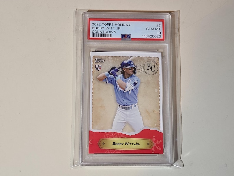 Op de afbeelding: Een honkbalkaart van Bobby Witt Jr. in een Kansas City Royals-uniform, beoordeeld en beschermd in een houder. De kaart heeft de tekst "2022 Topps Holiday" en "Bobby Witt Jr."