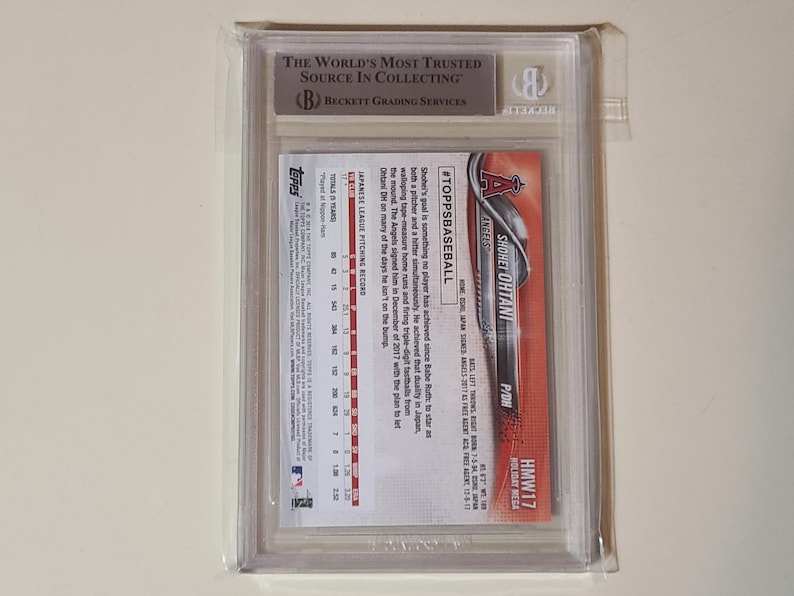 May include: A baseball card of Shohei Ohtani in a protective case. The card features the Topps logo and the Angels team logo. The case has a Beckett Grading Services label.