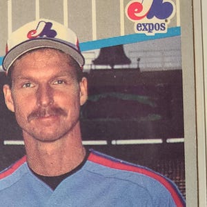 May include: A baseball card featuring a man wearing a baseball cap with the Expos logo. He is wearing a blue and red baseball jersey. The Expos logo is also visible on the top right corner of the card.