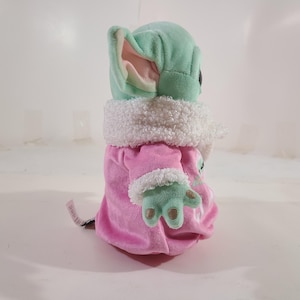 Pu&ograve; includere: Bambola di peluche di un personaggio con grandi orecchie e pelle verde, che indossa un cappotto rosa con finiture bianche. La bambola ha un colletto bianco e soffice e mani verdi. La bambola &egrave; alta circa 15 cm.