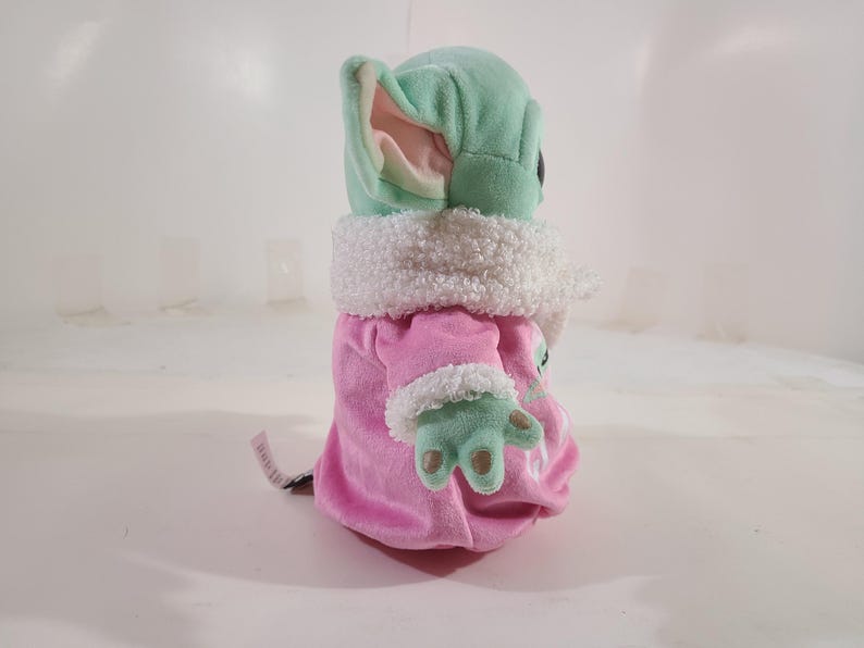 Pu&ograve; includere: Bambola di peluche di un personaggio con grandi orecchie e pelle verde, che indossa un cappotto rosa con finiture bianche. La bambola ha un colletto bianco e soffice e mani verdi. La bambola &egrave; alta circa 15 cm.