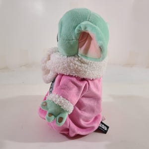 Pu&ograve; includere: Un peluche di un personaggio con una testa grande, grandi orecchie e pelle verde. Il personaggio indossa una veste rosa con finiture bianche e ha le mani verdi. Il giocattolo &egrave; un personaggio popolare di un franchise di fantascienza.