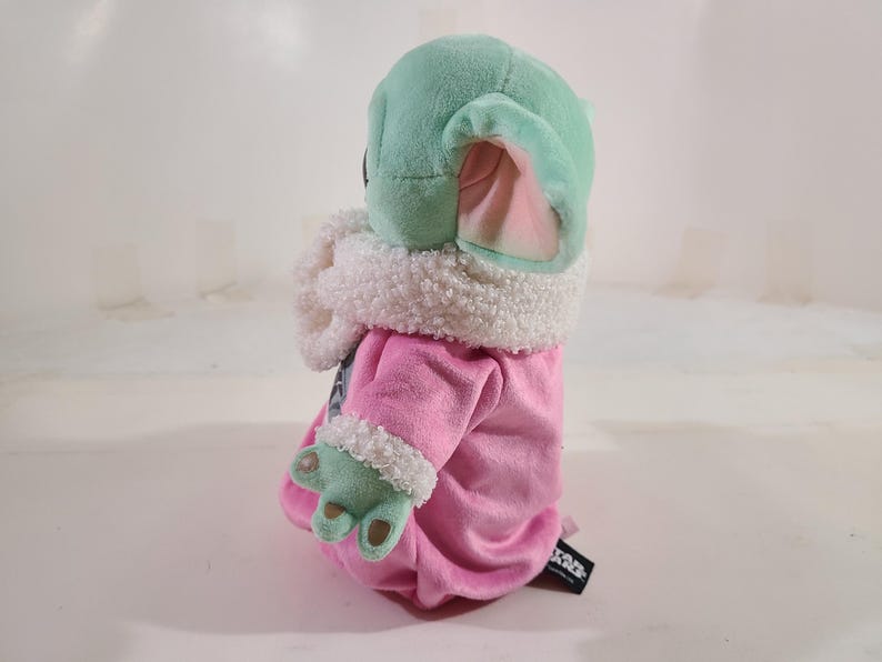 Pu&ograve; includere: Un peluche di un personaggio con una testa grande, grandi orecchie e pelle verde. Il personaggio indossa una veste rosa con finiture bianche e ha le mani verdi. Il giocattolo &egrave; un personaggio popolare di un franchise di fantascienza.
