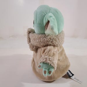 May include: A plush toy of a character with large ears, green skin, and a tan robe. The toy has a fluffy collar and cuffs. The character's hands are visible, and a tag is attached to the robe.