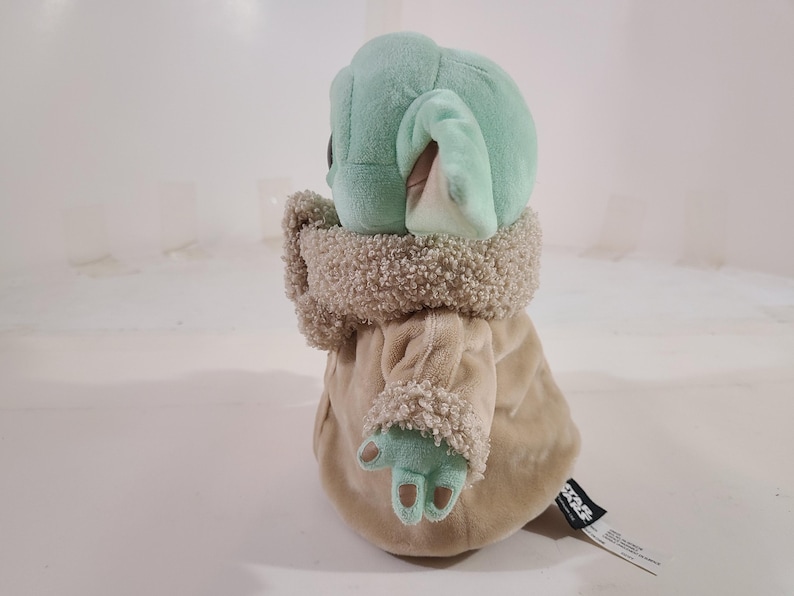 May include: A plush toy of a character with large ears, green skin, and a tan robe. The toy has a fluffy collar and cuffs. The character's hands are visible, and a tag is attached to the robe.