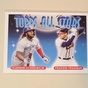 May include: A Topps All Stars baseball card featuring Vladimir Guerrero Jr. in a grey Toronto uniform and Freddie Freeman in a white Atlanta uniform. The background is a blue gradient with star accents. The names are printed in red at the bottom.