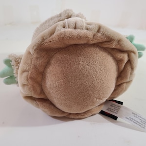 May include: A plush toy in the shape of a character with a large head and large ears. The toy is primarily tan with green accents on the hands. A tag is attached to the bottom.