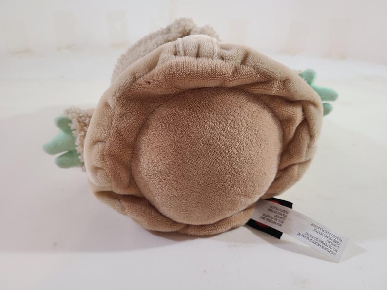 May include: A plush toy in the shape of a character with a large head and large ears. The toy is primarily tan with green accents on the hands. A tag is attached to the bottom.