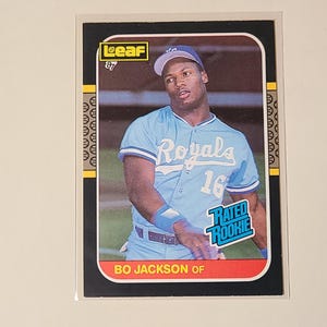 May include: A baseball card featuring Bo Jackson of the Kansas City Royals. The card has a black border with gold accents and a red bottom border. Jackson is wearing a light blue Royals jersey with the number 16.